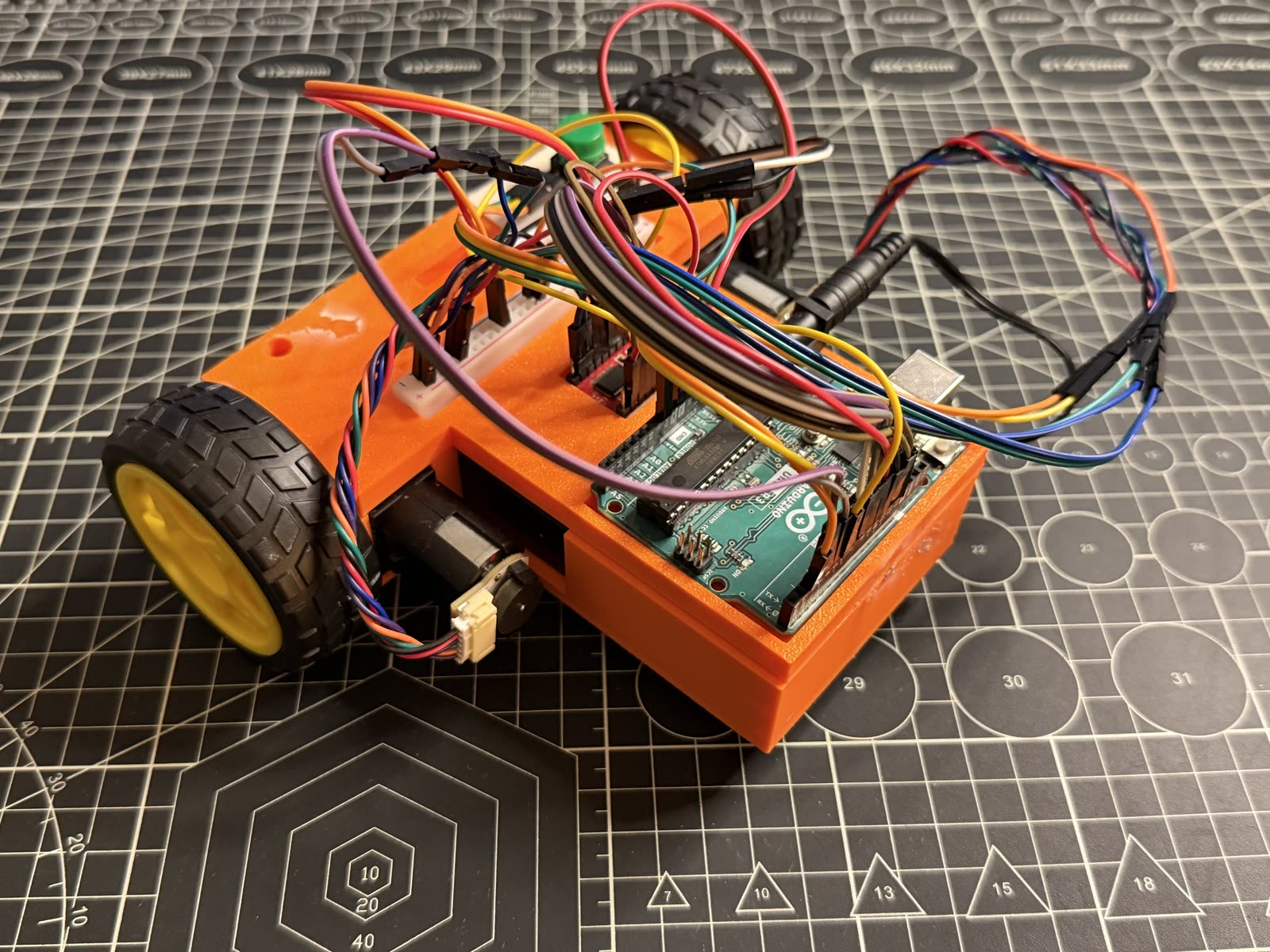 Compact orange 3D-printed electric vehicle with a genuine Arduino Uno, breadboard, and yellow-hub wheels on a cutting mat.
