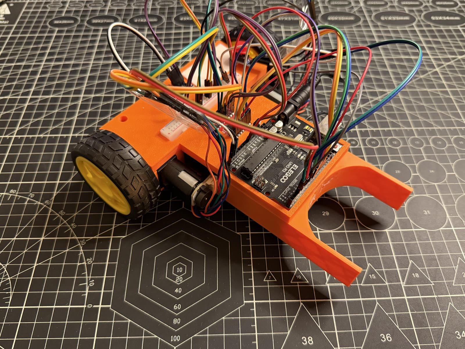 Orange 3D-printed robot car with a long front guide arm, an Elegoo Arduino Uno, breadboard, and a tangle of jumper wires on a cutting mat.